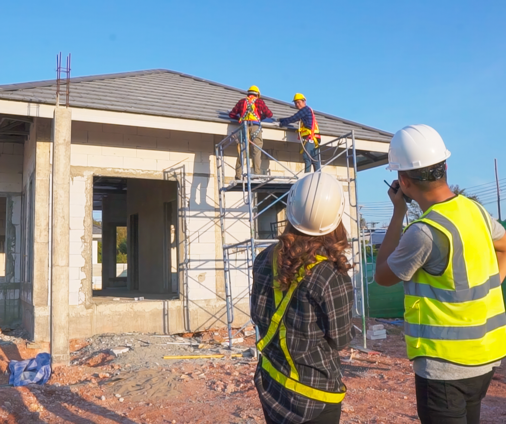 Two construction workers in safety gear discussing plans on a building site with scaffolding and a partially built house.