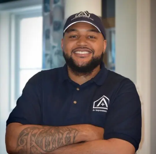 IMG_9231 (1) A smiling man with a beard wearing a navy blue polo shirt and cap, standing indoors with arms crossed.