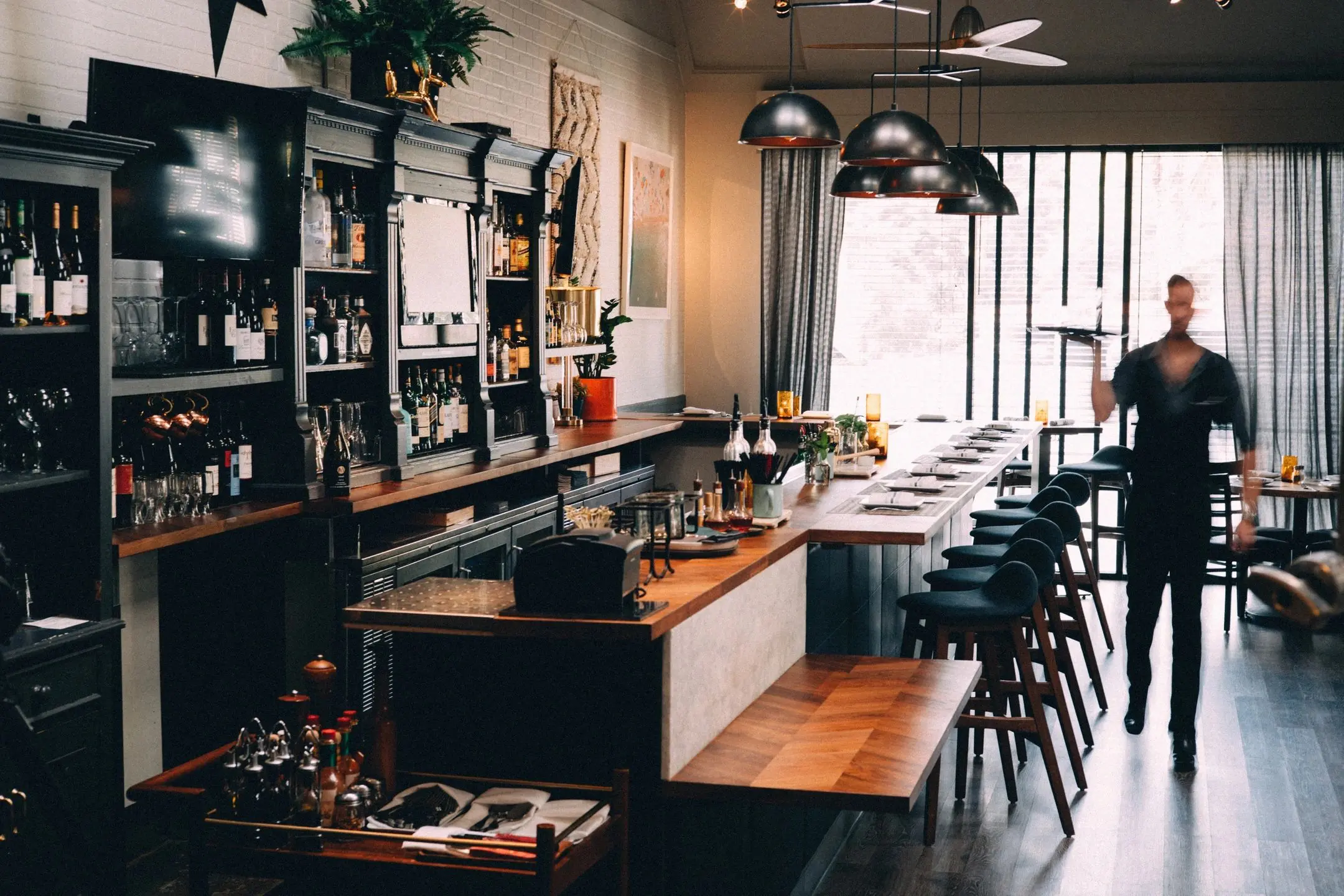 fm=f_uyrlZzzaWPF22Vnng Cozy cafe interior with wooden tables, chairs, and a bar counter under hanging pendant lights.