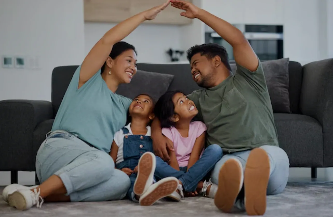 A happy family of four sitting on the floor, smiling and making heart shapes with their arms.