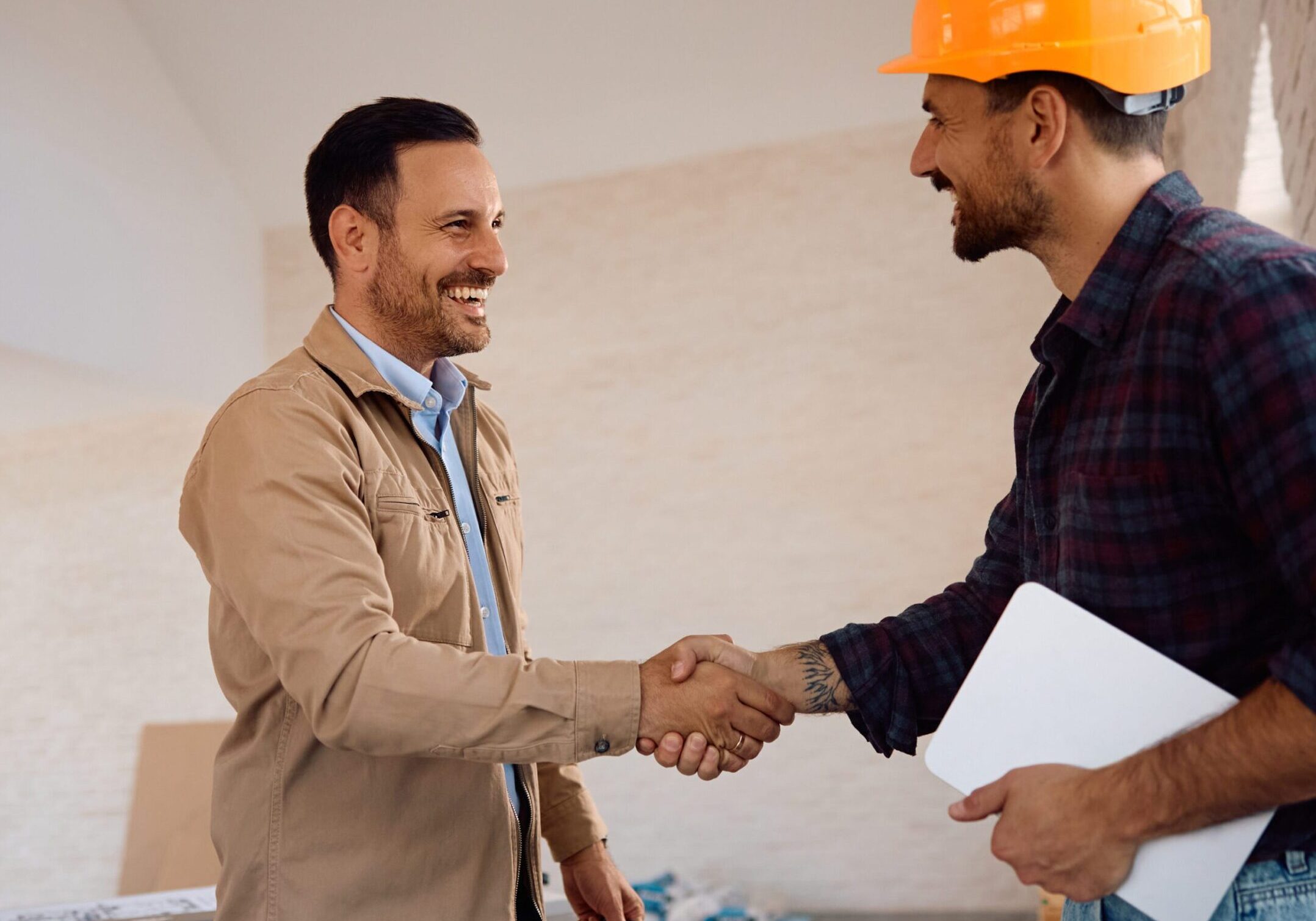 Two men shaking hands in a friendly manner, one wearing a safety helmet and holding documents, likely on a construction site.